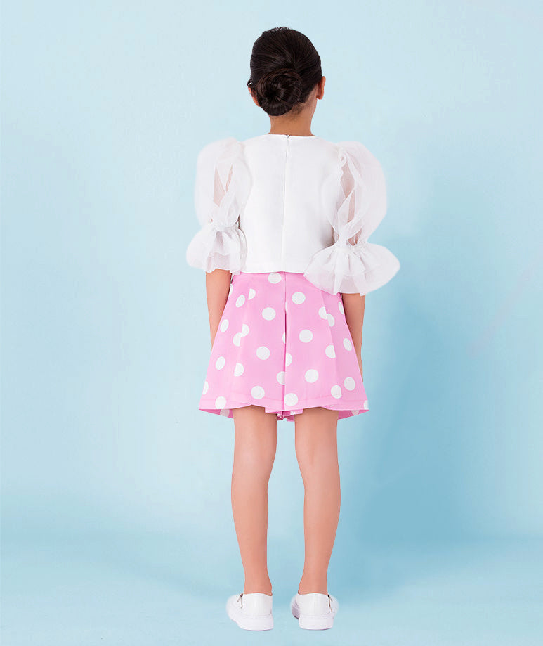 Girl wearing a white organza puff-sleeve blouse with ruffled cuffs paired with a pink pleated polka-dot skirt by Mama Luma. A playful and elegant outfit perfect for birthdays, summer parties, and spring celebrations. Available in girls’ sizes 3–12 years.
