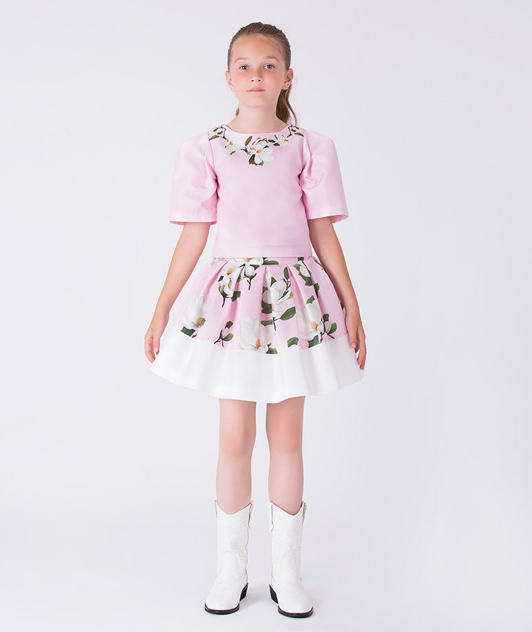 Front view of a pink floral girls occasion outfit featuring a short sleeve structured top and a matching flared skirt with contrast hem, styled for birthdays, weddings, garden parties, and special occasions, available in sizes 3–12