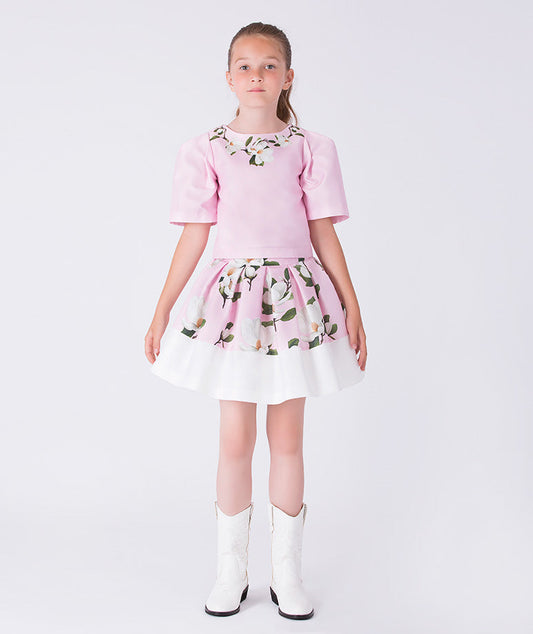 Front view of a pink floral girls occasion outfit featuring a short sleeve structured top and a matching flared skirt with contrast hem, styled for birthdays, weddings, garden parties, and special occasions, available in sizes 3–12