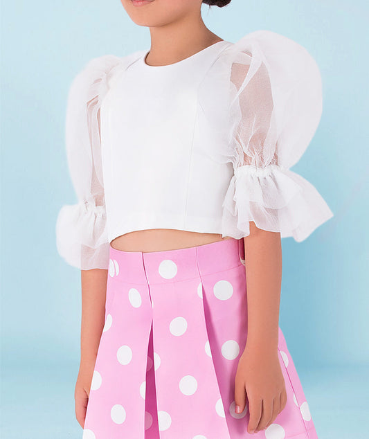 Girl wearing a white organza puff-sleeve blouse with ruffled cuffs paired with a pink pleated polka-dot skirt by Mama Luma. A playful and elegant outfit perfect for birthdays, summer parties, and spring celebrations. Available in girls’ sizes 3–12 years.
