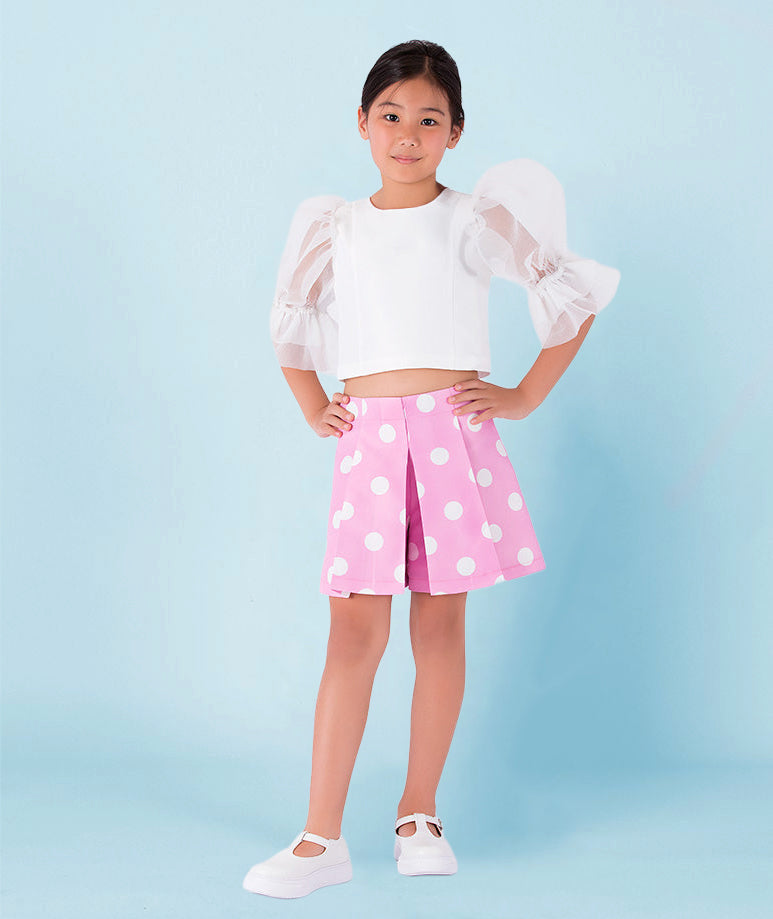 Girl wearing a white organza puff-sleeve blouse with ruffled cuffs paired with a pink pleated polka-dot skirt by Mama Luma. A playful and elegant outfit perfect for birthdays, summer parties, and spring celebrations. Available in girls’ sizes 3–12 years.