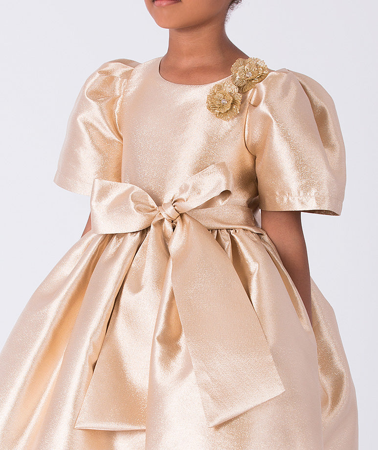 Close-up of gold satin girls dress featuring floral shoulder appliqué and oversized waist bow detail.